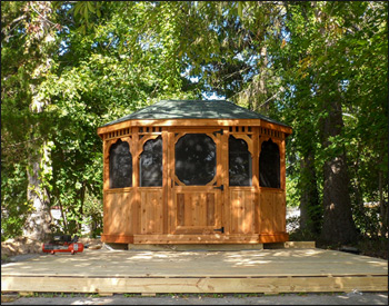 8 x 12 Cedar Oval Gazebo shown with Red Cedar Deck, Solid Bottom Railings, Straight Posts, Standard Braces, Top railing Sections, Straight Fascia, 4 Track Window System with Screens, Cedar Tongue and Groove Ceiling, Rustic Evergreen Asphalt Shingles, Cedar Tone Stain/Sealer, Appliance White Avruc Outdoor Ceiling Fan w/ Light, Stainless Steel Hardware, Screened Floor, Hidden Wiring w/ 1 Receptacle & Switch, Receptacle, and Bench Sections.